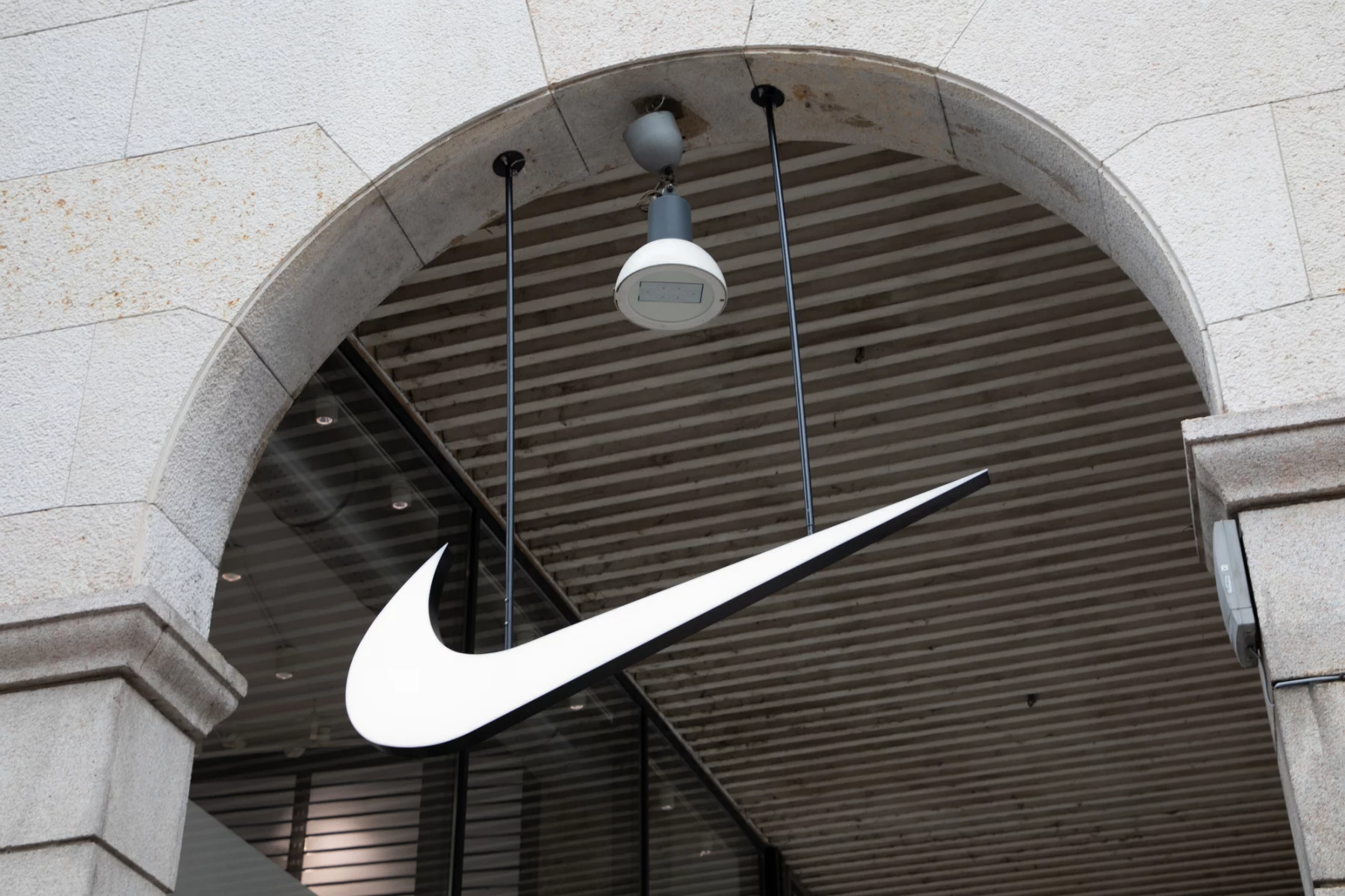 FootballR - NFL - Ein großes, weißes Nike Swoosh Logo hängt unter einem Steintorbogen vor einem Geschäft. Der Torbogen hat eine beige Fassade mit sichtbaren Steinstrukturen. Über dem Logo, das mit ihrer NFL-Partnerschaft in Verbindung steht, ist an der Decke, die mit horizontalen Holzlamellen versehen ist, eine einzelne weiße Überwachungskamera mit eingebauter Leuchte angebracht. Diese Beschreibung wurde automatisch generiert.
