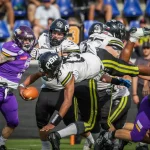 FootballR - NFL - Ein Footballspieler in weißem Trikot und schwarzer Hose hechtet mit dem Ball in der Hand über das Spielfeld und zielt auf einen Touchdown beim Austrian Bowl XXXIX. Er ist in der Luft und Verteidiger in lila Trikots versuchen, ihn zu tackeln. Andere Spieler in weißen und schwarzen Trikots blockieren die Verteidiger. Im Hintergrund sind Zuschauer auf blauen Sitzen zu sehen. Diese Beschreibung wurde automatisch generiert.