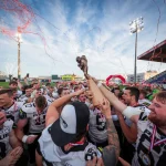 FootballR - NFL - Eine jubelnde Football-Mannschaft in weiß-schwarzen Trikots mit der Aufschrift „PREGLUE“ auf der Brust reckt unter einem klaren blauen Himmel gemeinsam den Pokal des Austrian Bowl XXXIX-Siegers in die Höhe. Rotes Konfetti und Luftschlangen liegen in der Luft, während sie auf einem gut beleuchteten Footballfeld feiern, im Hintergrund sind Zuschauerränge zu sehen. Diese Beschreibung wurde automatisch generiert.