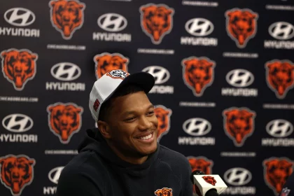 FootballR - NFL - Eine lächelnde Person, DJ Moore, die eine weiße Kappe und einen dunklen Kapuzenpullover mit einem Bärenlogo trägt, sitzt an einem Pressekonferenztisch. Dahinter ist ein Hintergrund voller Logos, darunter Hyundai und ein Bärenkopf, der mit dem NFL-Team Chicago Bears in Verbindung steht. DJ Moore spricht in das Mikrofon mit demselben Logo. Diese Beschreibung wurde automatisch generiert.