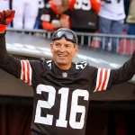 FootballR - NFL Bernie Kosar - Ein Mann in einem schwarzen Footballtrikot mit der Nummer 216, schwarzen und orangefarbenen Handschuhen und einer Schutzbrille streckt beide Arme zum Jubeln in die Höhe. Er steht in einem Stadion voller Fans, von denen einige Browns-Legende-Trikots tragen, die an Bernie Kosars Ära erinnern. Diese Beschreibung wurde automatisch generiert.