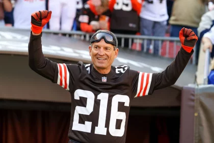 FootballR - NFL Bernie Kosar - Ein Mann in einem schwarzen Footballtrikot mit der Nummer 216, schwarzen und orangefarbenen Handschuhen und einer Schutzbrille streckt beide Arme zum Jubeln in die Höhe. Er steht in einem Stadion voller Fans, von denen einige Browns-Legende-Trikots tragen, die an Bernie Kosars Ära erinnern. Diese Beschreibung wurde automatisch generiert.