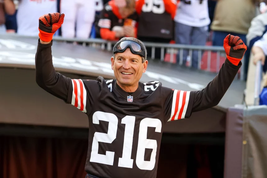 FootballR - NFL Bernie Kosar - Ein Mann in einem schwarzen Footballtrikot mit der Nummer 216, schwarzen und orangefarbenen Handschuhen und einer Schutzbrille streckt beide Arme zum Jubeln in die Höhe. Er steht in einem Stadion voller Fans, von denen einige Browns-Legende-Trikots tragen, die an Bernie Kosars Ära erinnern. Diese Beschreibung wurde automatisch generiert.