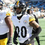 FootballR - NFL - Cameron Sutton - Zwei American-Football-Spieler der Pittsburgh Steelers stehen auf dem Spielfeld. Der Spieler im Vordergrund, Cameron Sutton, trägt ein weiß-gelbes Trikot mit der Nummer 20, während der Spieler hinter ihm die Nummer 72 trägt. Sie stehen auf einem Footballfeld mit einer verschwommenen Stadionmenge im Hintergrund. Diese Beschreibung wurde automatisch generiert.