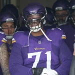 FootballR - NFL - Eine Gruppe von Footballspielern in Minnesota-Vikings-Uniformen steht zusammen in einem Tunnel und bereitet sich darauf vor, das Spielfeld zu betreten. Der Spieler in der Mitte, Christian Darrisaw, bestbezahlter Offensive Tackle mit einem Mega-Vertrag, lächelt breit. Alle Spieler tragen lila Helme und Uniformen mit goldenen Akzenten. Diese Beschreibung wurde automatisch generiert.