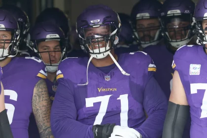 FootballR - NFL - Eine Gruppe von Footballspielern in Minnesota-Vikings-Uniformen steht zusammen in einem Tunnel und bereitet sich darauf vor, das Spielfeld zu betreten. Der Spieler in der Mitte, Christian Darrisaw, bestbezahlter Offensive Tackle mit einem Mega-Vertrag, lächelt breit. Alle Spieler tragen lila Helme und Uniformen mit goldenen Akzenten. Diese Beschreibung wurde automatisch generiert.