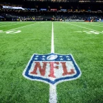 FootballR - NFL Saison 2024 - Ein Footballfeld mit dem NFL-Logo in der Mitte zwischen der 20- und 30-Yard-Linie. Das Stadion ist beleuchtet und im Hintergrund voller Zuschauer. Eine Anzeigetafel zeigt „FINAL STATS“ für die Saison 2023 an. Das Feld hat deutlich erkennbare weiße Yard-Linien und Hash-Markierungen, was auf eine gut gepflegte Spielfläche hinweist. Diese Beschreibung wurde automatisch generiert.