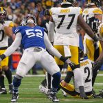 FootballR - NFL - Samson Ebukam, in seiner blauen Uniform mit der Nummer 52, steht mit ausgestreckten Armen und nach hinten geneigtem Kopf da, um zu jubeln. Spieler in weißen und gelben Uniformen reagieren auf das Spiel, einige knien oder stehen. Das Spielfeld ist mit weißen Linien markiert, während die Menge als verschwommener Hintergrund bleibt. Diese Beschreibung wurde automatisch generiert.