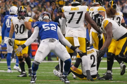 FootballR - NFL - Samson Ebukam, in seiner blauen Uniform mit der Nummer 52, steht mit ausgestreckten Armen und nach hinten geneigtem Kopf da, um zu jubeln. Spieler in weißen und gelben Uniformen reagieren auf das Spiel, einige knien oder stehen. Das Spielfeld ist mit weißen Linien markiert, während die Menge als verschwommener Hintergrund bleibt. Diese Beschreibung wurde automatisch generiert.