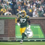 FootballR - NFL Kenny Clark - Ein Footballspieler der Green Bay Packers in grün-gelber Uniform mit der Nummer 97 rennt auf das Spielfeld. Der Defense Star steht vor einem Hintergrund jubelnder Fans, von denen viele Packers-Kleidung tragen. Auf der Backsteinmauer im Hintergrund ist ein großes „G“-Logo zu sehen. Diese Beschreibung wurde automatisch generiert.