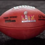 FootballR und die NFL Playoffs - Around the NFL - Eine Nahaufnahme eines offiziellen Wilson NFL-Footballs auf einer dunklen Oberfläche. Der Football zeigt in der Mitte das Super Bowl-Logo, links „THE DUKE“ und rechts die Unterschrift des Commissioners. Die Nähte sind oben auf dem Ball sichtbar, ein perfektes Thema für eine Diskussion im Podcast „Around the NFL“. Diese Beschreibung wurde automatisch generiert.