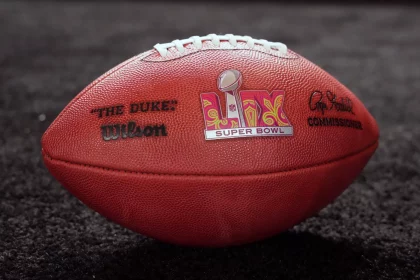 FootballR und die NFL Playoffs - Around the NFL - Eine Nahaufnahme eines offiziellen Wilson NFL-Footballs auf einer dunklen Oberfläche. Der Football zeigt in der Mitte das Super Bowl-Logo, links „THE DUKE“ und rechts die Unterschrift des Commissioners. Die Nähte sind oben auf dem Ball sichtbar, ein perfektes Thema für eine Diskussion im Podcast „Around the NFL“. Diese Beschreibung wurde automatisch generiert.