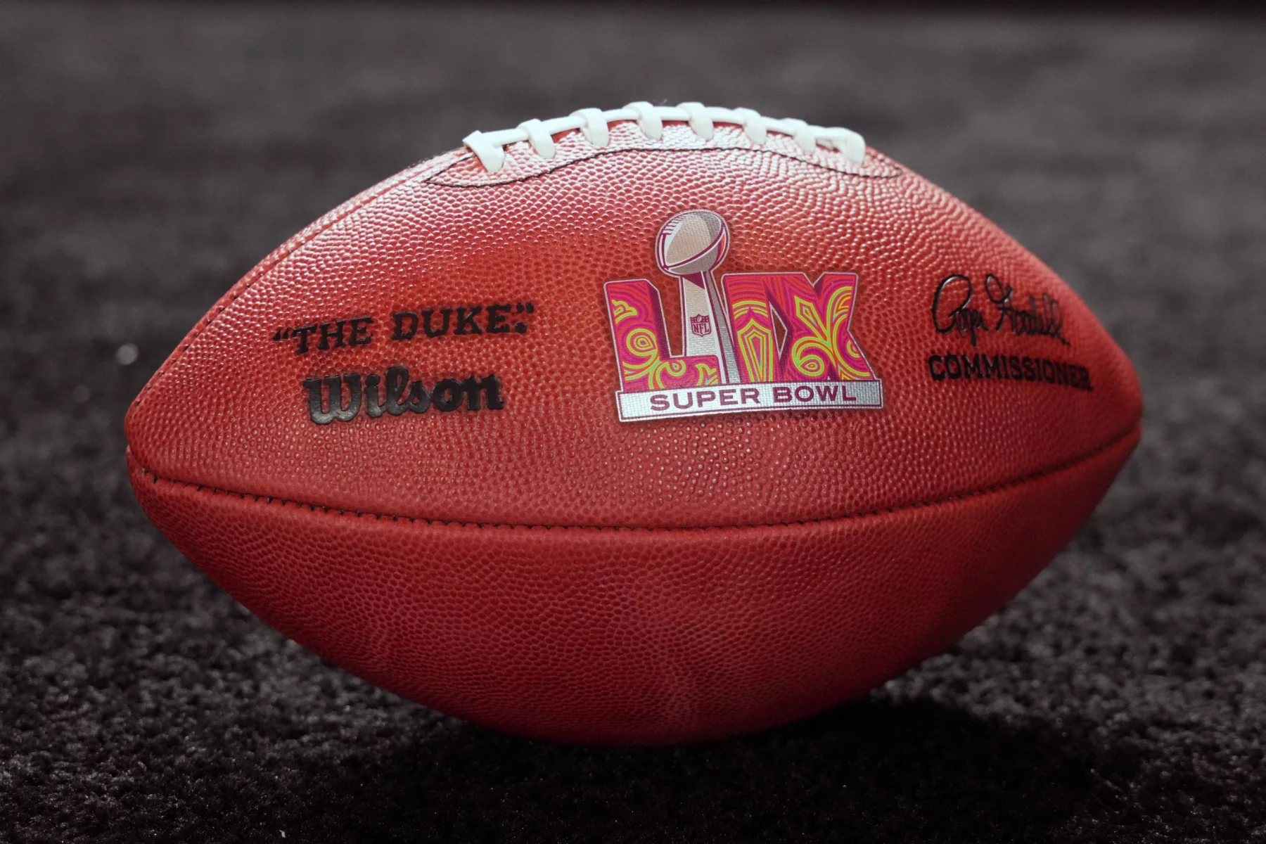 FootballR und die NFL Playoffs - Around the NFL - Eine Nahaufnahme eines offiziellen Wilson NFL-Footballs auf einer dunklen Oberfläche. Der Football zeigt in der Mitte das Super Bowl-Logo, links „THE DUKE“ und rechts die Unterschrift des Commissioners. Die Nähte sind oben auf dem Ball sichtbar, ein perfektes Thema für eine Diskussion im Podcast „Around the NFL“. Diese Beschreibung wurde automatisch generiert.