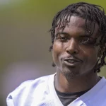 FootballR - NFL - Khyree Jackson - Eine Person mit geflochtenen Haaren ist von der Brust aufwärts zu sehen und lächelt leicht, während sie in die Kamera blickt. Sie trägt ein weißes Sporttrikot mit einem schwarzen Hemd darunter. Der Hintergrund ist verschwommen grün und lässt eine Szene im Freien vermuten. Die Szene ruft Bilder von Vikings-Neuling Khyree Jackson an seinem freien Tag hervor. Diese Beschreibung wurde automatisch generiert.