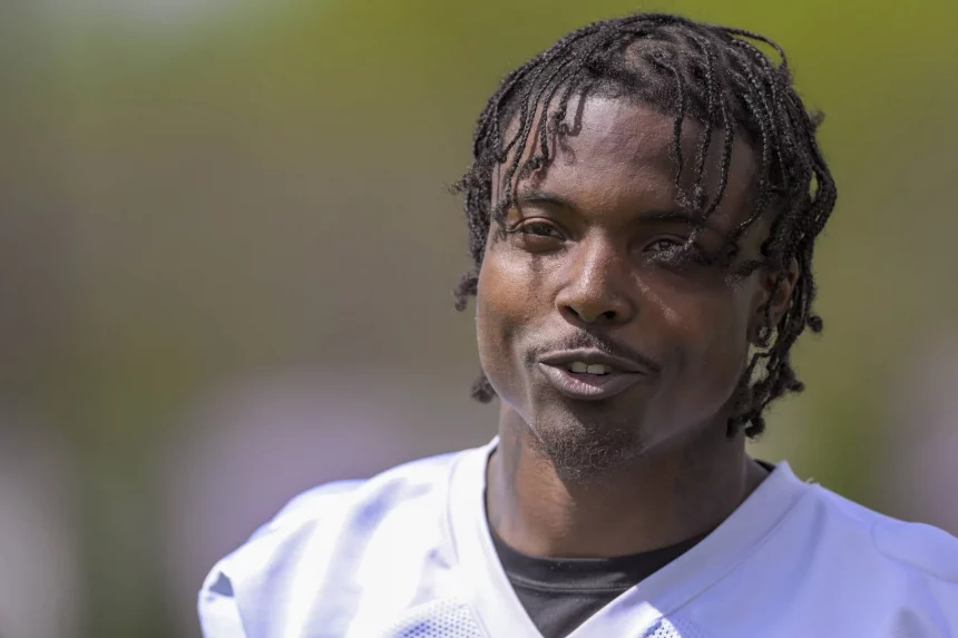 FootballR - NFL - Khyree Jackson - Eine Person mit geflochtenen Haaren ist von der Brust aufwärts zu sehen und lächelt leicht, während sie in die Kamera blickt. Sie trägt ein weißes Sporttrikot mit einem schwarzen Hemd darunter. Der Hintergrund ist verschwommen grün und lässt eine Szene im Freien vermuten. Die Szene ruft Bilder von Vikings-Neuling Khyree Jackson an seinem freien Tag hervor. Diese Beschreibung wurde automatisch generiert.