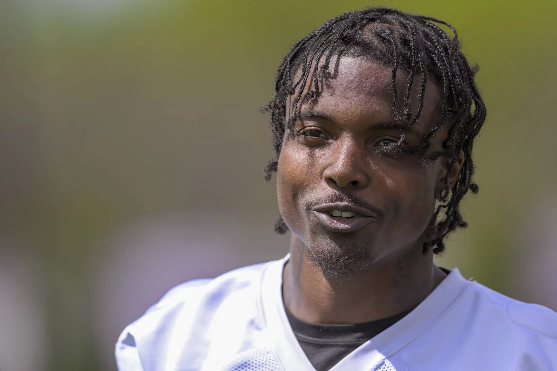 FootballR - NFL - Khyree Jackson - Eine Person mit geflochtenen Haaren ist von der Brust aufwärts zu sehen und lächelt leicht, während sie in die Kamera blickt. Sie trägt ein weißes Sporttrikot mit einem schwarzen Hemd darunter. Der Hintergrund ist verschwommen grün und lässt eine Szene im Freien vermuten. Die Szene ruft Bilder von Vikings-Neuling Khyree Jackson an seinem freien Tag hervor. Diese Beschreibung wurde automatisch generiert.