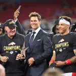 FootballR - NFL - Ein Mann im dunklen Anzug (Tom Brady) steht lächelnd mit einer Trophäe da, flankiert von zwei Personen in schwarzen „Champions“-Shirts und Kappen, darunter Adrian Martinez. Die Gruppe feiert auf dem Stadionfeld, hinter ihnen stehen mehrere jubelnde Fans der Birmingham Stallions. Die Leute machen Fotos und jubeln, im Hintergrund sind die Stadionsitze zu sehen. Diese Beschreibung wurde automatisch generiert.