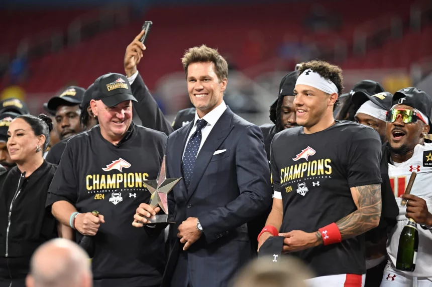 FootballR - NFL - Ein Mann im dunklen Anzug (Tom Brady) steht lächelnd mit einer Trophäe da, flankiert von zwei Personen in schwarzen „Champions“-Shirts und Kappen, darunter Adrian Martinez. Die Gruppe feiert auf dem Stadionfeld, hinter ihnen stehen mehrere jubelnde Fans der Birmingham Stallions. Die Leute machen Fotos und jubeln, im Hintergrund sind die Stadionsitze zu sehen. Diese Beschreibung wurde automatisch generiert.