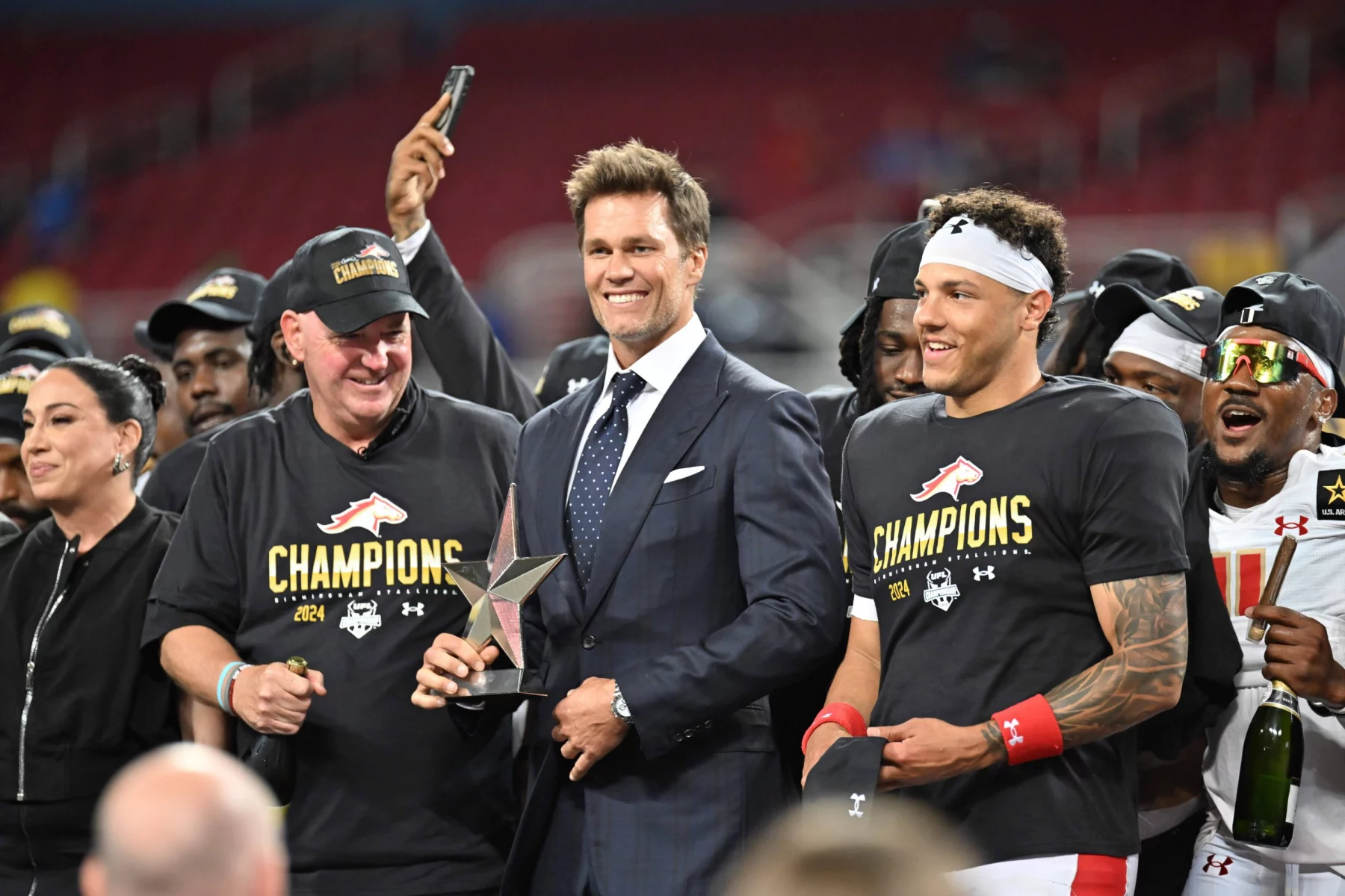 FootballR - NFL - Ein Mann im dunklen Anzug (Tom Brady) steht lächelnd mit einer Trophäe da, flankiert von zwei Personen in schwarzen „Champions“-Shirts und Kappen, darunter Adrian Martinez. Die Gruppe feiert auf dem Stadionfeld, hinter ihnen stehen mehrere jubelnde Fans der Birmingham Stallions. Die Leute machen Fotos und jubeln, im Hintergrund sind die Stadionsitze zu sehen. Diese Beschreibung wurde automatisch generiert.