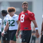 FootballR - NFL - Aaron Rodgers in einem roten Trainings-Footballtrikot mit der Nummer 8 steht auf einem Feld. Im Hintergrund sind andere Spieler zu sehen, die weiße Trikots mit grünen Nummern tragen, entweder stehend oder gehend. Der Mann ähnelt Aaron Rodgers beim Training, blickt leicht nach links und in der Ferne ist ein Gebäude zu sehen. Diese Beschreibung wurde automatisch generiert.