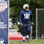 FootballR - NFL Christian Barmore - Ein Footballspieler, der ein marineblaues Patriots-Trikot mit der Nummer 90, einen weißen Helm und einen roten Gesichtsschutz trägt, läuft auf einer Rasenfläche in der Nähe eines Maschendrahtzauns. Der Spieler ist Teil eines Trainingslagers, was durch ein Banner mit den Aufschriften „Trainingslager 2024“ und „Mass General Brigham“ gekennzeichnet ist. Im Hintergrund sind Bäume zu sehen. Diese Beschreibung wurde automatisch generiert.