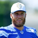 FootballR - NFL - Cooper Kupp, ein Rams-Star, lächelt, während er eine weiße Kappe mit einem Widderkopf-Logo und ein blaues Trikot trägt. Das Trikot zeigt ein Nike-Logo auf der rechten Schulter und das Teamlogo auf der Brust. Vor einem verschwommenen Hintergrund aus grünem Laub zeigt Kupp seinen Vollbart und blickt leicht zur Seite. Diese Beschreibung wurde automatisch generiert.