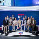 FootballR - NFL - Eine Gruppe von elf Personen, zehn Männer und eine Frau, posiert in einem professionellen RTL NFL Studio vor einem blau-weißen NFL Live-Hintergrund. Sie tragen legere Business-Kleidung und wirken gelassen und bereit zum Handeln – einige sitzen auf einer Bank, andere stehen dahinter. Vor ihnen liegt ein Offensiv-Football auf dem Boden. Diese Beschreibung wurde automatisch generiert.