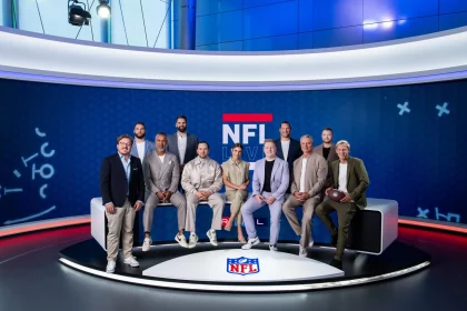 FootballR - NFL - Eine Gruppe von elf Personen, zehn Männer und eine Frau, posiert in einem professionellen RTL NFL Studio vor einem blau-weißen NFL Live-Hintergrund. Sie tragen legere Business-Kleidung und wirken gelassen und bereit zum Handeln – einige sitzen auf einer Bank, andere stehen dahinter. Vor ihnen liegt ein Offensiv-Football auf dem Boden. Diese Beschreibung wurde automatisch generiert.