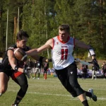 FootballR - NFL - Zwei männliche Flag Football-Spieler sind in Aktion. Der Spieler links in schwarzer Uniform rennt mit dem Ball, während ihn der Spieler rechts in weiß-roter Uniform mit der Nummer 11 verfolgt. Im Hintergrund sind Bäume und Zuschauer beim Flag Football WM Finale in Österreich zu sehen. Diese Beschreibung wurde automatisch generiert.