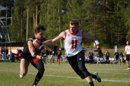 FootballR - NFL - Zwei männliche Flag Football-Spieler sind in Aktion. Der Spieler links in schwarzer Uniform rennt mit dem Ball, während ihn der Spieler rechts in weiß-roter Uniform mit der Nummer 11 verfolgt. Im Hintergrund sind Bäume und Zuschauer beim Flag Football WM Finale in Österreich zu sehen. Diese Beschreibung wurde automatisch generiert.