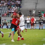 FootballR - Flag Football -Weltmeisterschaft - Ein Footballspieler in einem roten Trikot mit der Nummer 6 und schwarzen Shorts versucht, einem Tackling eines Spielers in weißem Trikot und Shorts auf dem Feld auszuweichen. Ein weiterer Spieler in einem weißen Trikot folgt ihm dicht auf den Fersen. Im Hintergrund sind Zuschauer, die mexikanische Flagge und ein Österreich-Banner zu sehen, das prominent auf der Tribüne zu sehen ist. Diese Beschreibung wurde automatisch generiert.