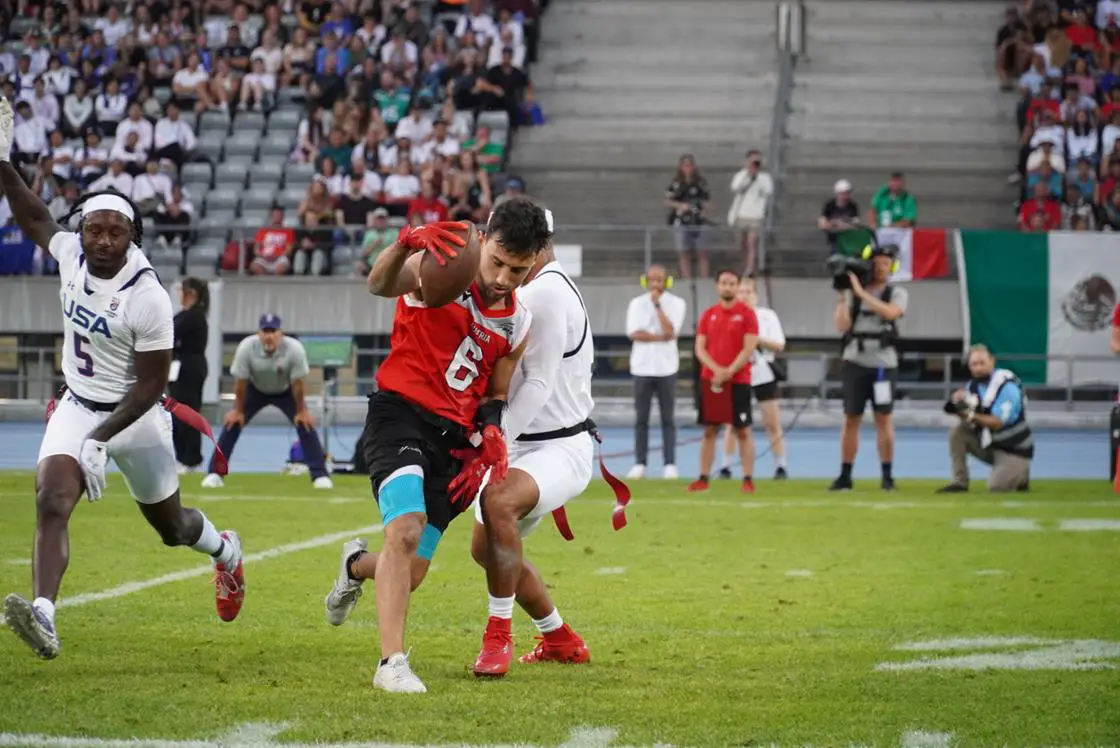 FootballR - Flag Football -Weltmeisterschaft - Ein Footballspieler in einem roten Trikot mit der Nummer 6 und schwarzen Shorts versucht, einem Tackling eines Spielers in weißem Trikot und Shorts auf dem Feld auszuweichen. Ein weiterer Spieler in einem weißen Trikot folgt ihm dicht auf den Fersen. Im Hintergrund sind Zuschauer, die mexikanische Flagge und ein Österreich-Banner zu sehen, das prominent auf der Tribüne zu sehen ist. Diese Beschreibung wurde automatisch generiert.