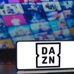 FootballR - European League of Football - NFL Game Pass 2024 - Ein Smartphone mit dem DAZN-Logo steht aufrecht auf einer teilweise sichtbaren Laptop-Tastatur. Im Hintergrund deutet ein unscharfer Bildschirm mit bunten Vorschaubildern auf eine Reihe von Medieninhalten hin, darunter Football-Erlebnis und NFL Game Pass 2024, die zum Schnäppchenpreis erhältlich sind. Diese Beschreibung wurde automatisch generiert.