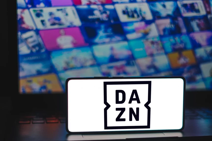 FootballR - European League of Football - NFL Game Pass 2024 - Ein Smartphone mit dem DAZN-Logo steht aufrecht auf einer teilweise sichtbaren Laptop-Tastatur. Im Hintergrund deutet ein unscharfer Bildschirm mit bunten Vorschaubildern auf eine Reihe von Medieninhalten hin, darunter Football-Erlebnis und NFL Game Pass 2024, die zum Schnäppchenpreis erhältlich sind. Diese Beschreibung wurde automatisch generiert.