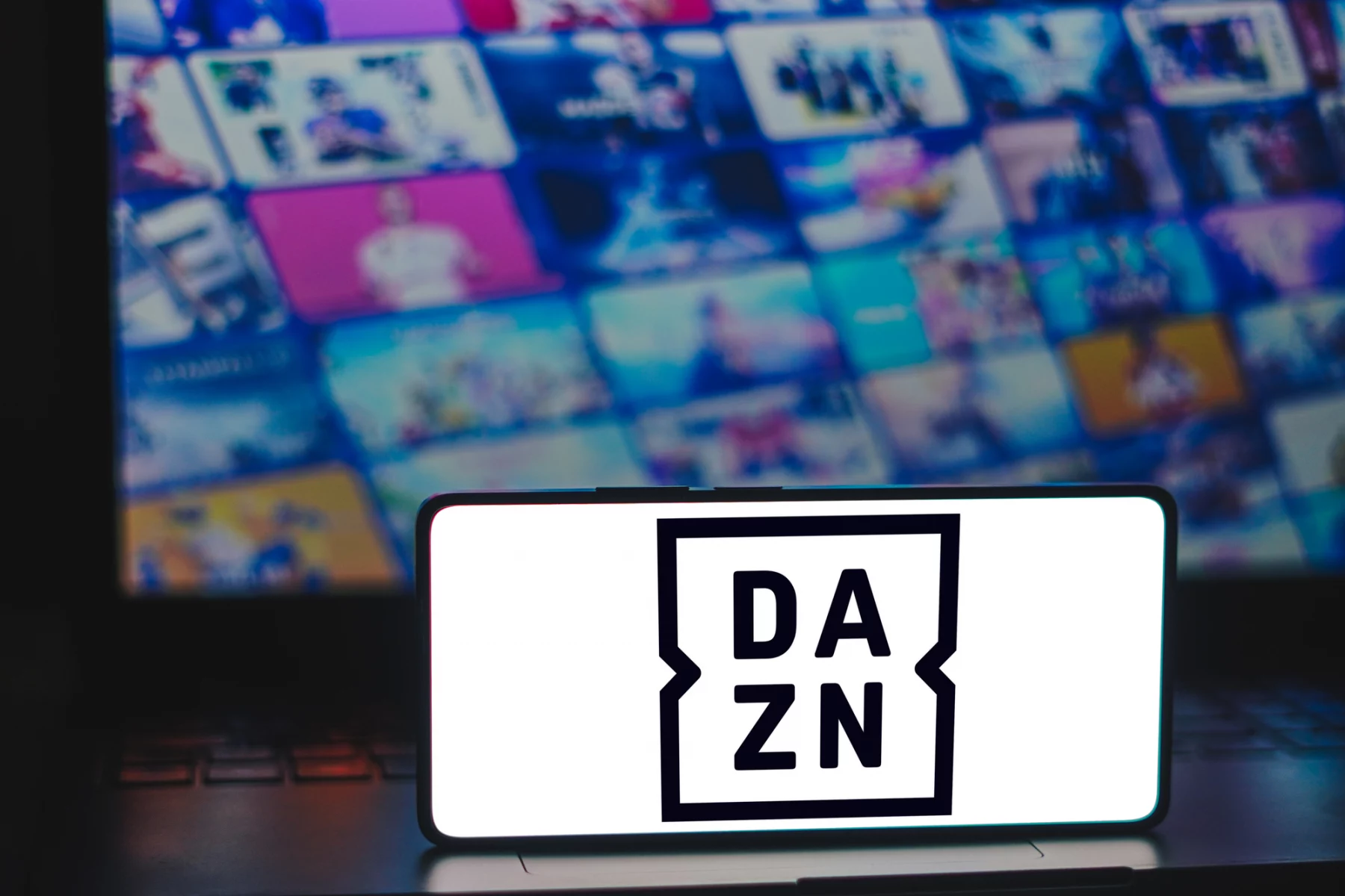 FootballR - European League of Football - NFL Game Pass 2024 - Ein Smartphone mit dem DAZN-Logo steht aufrecht auf einer teilweise sichtbaren Laptop-Tastatur. Im Hintergrund deutet ein unscharfer Bildschirm mit bunten Vorschaubildern auf eine Reihe von Medieninhalten hin, darunter Football-Erlebnis und NFL Game Pass 2024, die zum Schnäppchenpreis erhältlich sind. Diese Beschreibung wurde automatisch generiert.