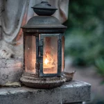 FootballR - NFL - Eine verwitterte Laterne mit Glasscheiben strahlt ein warmes, leuchtendes Licht aus. Sie steht auf einer Steinfläche neben einer Statue, im Hintergrund ist ein leicht verschwommener grün-weißer Garten zu sehen. Um den Sockel der Laterne herum sind kleine Steine und ein abgefallenes Blatt verstreut, was an Duane Thomas ruhige Stärke erinnert. Diese Beschreibung wurde automatisch generiert.