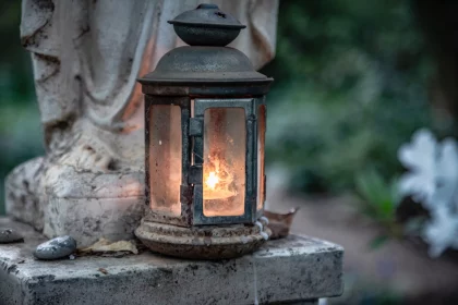 FootballR - NFL - Eine verwitterte Laterne mit Glasscheiben strahlt ein warmes, leuchtendes Licht aus. Sie steht auf einer Steinfläche neben einer Statue, im Hintergrund ist ein leicht verschwommener grün-weißer Garten zu sehen. Um den Sockel der Laterne herum sind kleine Steine und ein abgefallenes Blatt verstreut, was an Duane Thomas ruhige Stärke erinnert. Diese Beschreibung wurde automatisch generiert.