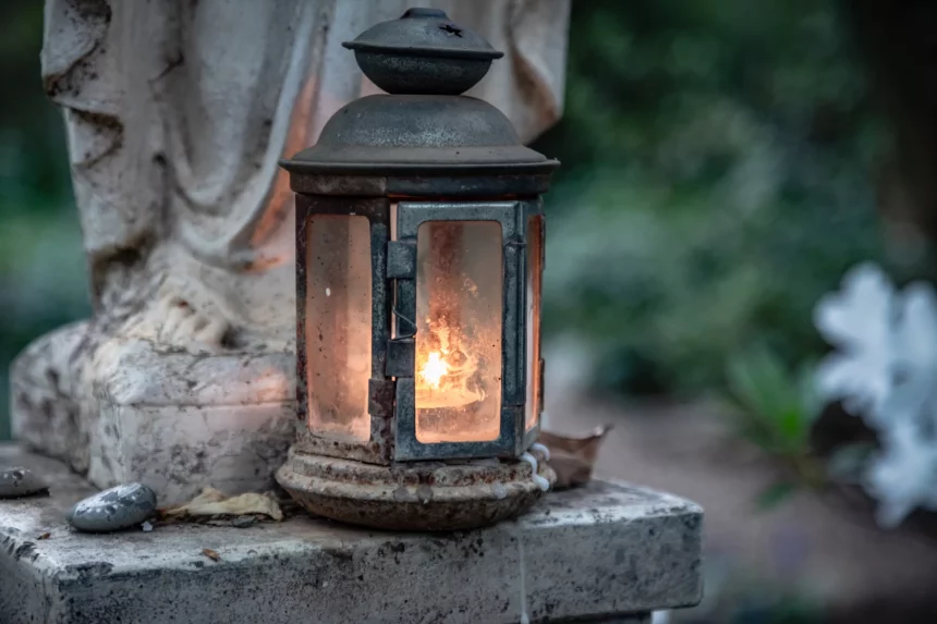 FootballR - NFL - Eine verwitterte Laterne mit Glasscheiben strahlt ein warmes, leuchtendes Licht aus. Sie steht auf einer Steinfläche neben einer Statue, im Hintergrund ist ein leicht verschwommener grün-weißer Garten zu sehen. Um den Sockel der Laterne herum sind kleine Steine und ein abgefallenes Blatt verstreut, was an Duane Thomas ruhige Stärke erinnert. Diese Beschreibung wurde automatisch generiert.