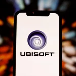 FootballR - NFL Primetime Fantasy - Ein Smartphone mit dem Ubisoft-Logo auf dem Bildschirm. Das Logo, ein blaues Spiraldesign mit der Aufschrift „UBISOFT“ darunter, fällt deutlich auf. Der Hintergrund ist mit mehrfarbigen Lichtstreifen verschwommen, was einen dynamischen und lebendigen Effekt rund um das Telefon erzeugt, der an ein intensives Live-Action-Fantasy-Spiel der NFL Primetime erinnert. Diese Beschreibung wurde automatisch generiert.