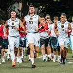 FootballR - NFL - Eine Gruppe männlicher Athleten in weißen Trainingsuniformen mit schwarzen und goldenen Akzenten läuft bei den IFAF Flag Football Weltmeisterschaften auf einem Feld. Der Athlet in der Mitte trägt die Nummer 23, flankiert von den Nummern 22 und 13. Hinter ihnen folgen weitere Teamkollegen in Rot und Schwarz. Im Hintergrund sind Bäume zu sehen. Diese Beschreibung wurde automatisch generiert.