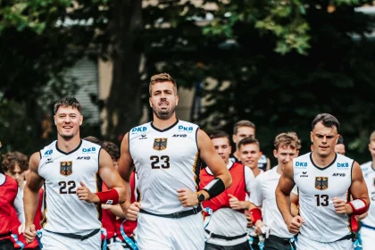 FootballR - NFL - Eine Gruppe männlicher Athleten in weißen Trainingsuniformen mit schwarzen und goldenen Akzenten läuft bei den IFAF Flag Football Weltmeisterschaften auf einem Feld. Der Athlet in der Mitte trägt die Nummer 23, flankiert von den Nummern 22 und 13. Hinter ihnen folgen weitere Teamkollegen in Rot und Schwarz. Im Hintergrund sind Bäume zu sehen. Diese Beschreibung wurde automatisch generiert.