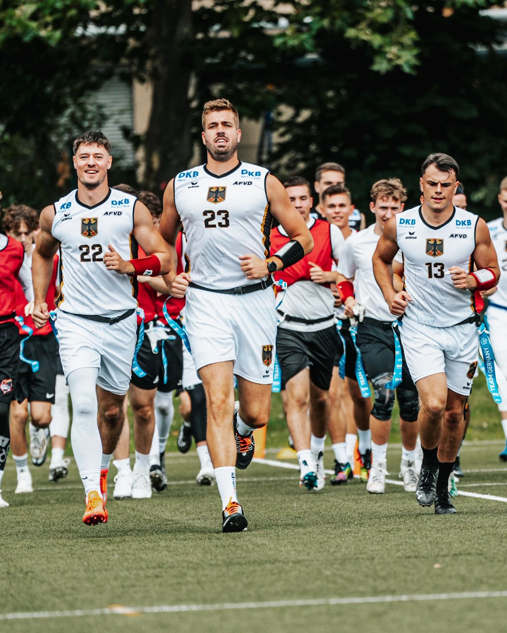 FootballR - NFL - Eine Gruppe männlicher Athleten in weißen Trainingsuniformen mit schwarzen und goldenen Akzenten läuft bei den IFAF Flag Football Weltmeisterschaften auf einem Feld. Der Athlet in der Mitte trägt die Nummer 23, flankiert von den Nummern 22 und 13. Hinter ihnen folgen weitere Teamkollegen in Rot und Schwarz. Im Hintergrund sind Bäume zu sehen. Diese Beschreibung wurde automatisch generiert.
