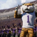 FootballR - NFL College Football 26 - Ein Tiger-Maskottchen, das triumphierend auf das College-Footballfeld zurückkehrt, trägt ein weißes Trikot mit der Nummer 1 und „LSU“ und steht mit beiden Zeigefingern nach oben auf einem Footballfeld. Im Hintergrund ist ein überfülltes Stadion mit Fans in Lila und Gold sowie einer Blaskapelle in passender lila Kleidung zu sehen. Diese Beschreibung wurde automatisch generiert.