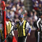 FootballR - NFL Hawk Eye Technologie - Schiedsrichter an der Seitenlinie eines Stadions während eines NFL-Spiels. Ein Schiedsrichter hält einen Downmarker und trägt gelbe Westen über seiner Uniform. Im Hintergrund ist eine verschwommene Menschenmenge zu sehen, die größtenteils in Lila gekleidet ist. Das Spielfeld und die Tribünen sind sichtbar, was auf ein Freiluftspiel schließen lässt. Diese Beschreibung wurde automatisch generiert.