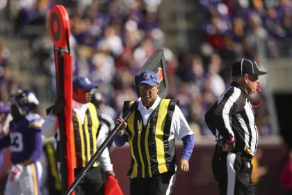 FootballR - NFL Hawk Eye Technologie - Schiedsrichter an der Seitenlinie eines Stadions während eines NFL-Spiels. Ein Schiedsrichter hält einen Downmarker und trägt gelbe Westen über seiner Uniform. Im Hintergrund ist eine verschwommene Menschenmenge zu sehen, die größtenteils in Lila gekleidet ist. Das Spielfeld und die Tribünen sind sichtbar, was auf ein Freiluftspiel schließen lässt. Diese Beschreibung wurde automatisch generiert.