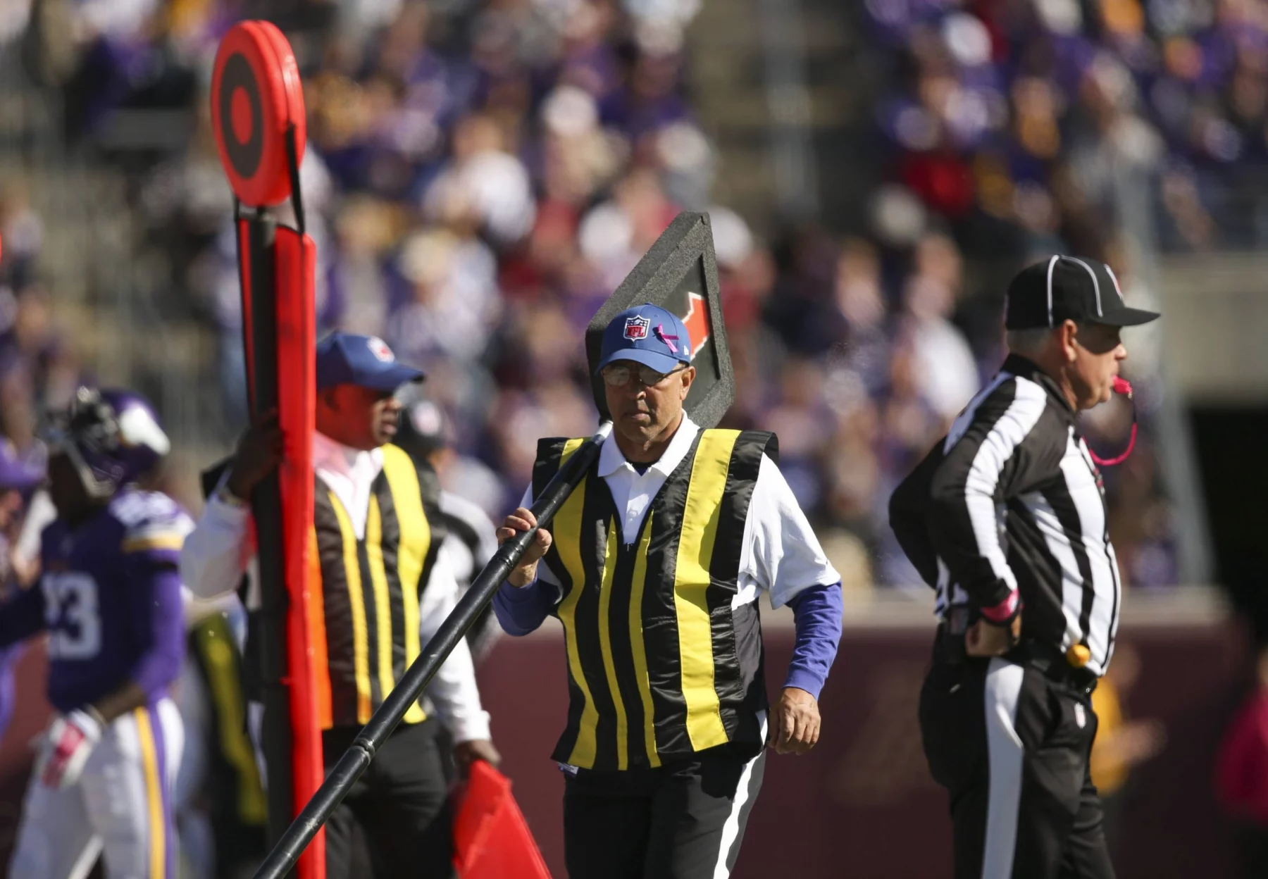 FootballR - NFL Hawk Eye Technologie - Schiedsrichter an der Seitenlinie eines Stadions während eines NFL-Spiels. Ein Schiedsrichter hält einen Downmarker und trägt gelbe Westen über seiner Uniform. Im Hintergrund ist eine verschwommene Menschenmenge zu sehen, die größtenteils in Lila gekleidet ist. Das Spielfeld und die Tribünen sind sichtbar, was auf ein Freiluftspiel schließen lässt. Diese Beschreibung wurde automatisch generiert.