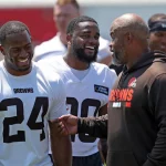FootballR - NFL - Drei Männer in Cleveland Browns-Ausrüstung unterhalten sich im Freien angeregt. Zwei Männer in weißen Sporttrikots mit den Nummern 24 und 30 lächeln, während sie einem kahlköpfigen Mann in einer braunen Jacke mit der orangefarbenen Aufschrift „Browns“ zuhören, der mit der Hand gestikuliert. Er freut sich sichtlich auf den Saisonstart mit Nick Chubb! Andere Personen sind im Hintergrund verschwommen. Diese Beschreibung wurde automatisch generiert.