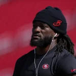 FootballR - NFL - Ein Mann mit langen Dreadlocks, einer schwarzen Beanie-Mütze mit rotem „B“-Logo und einem schwarzen Nike-T-Shirt mit dem 49ers-Star Brandon Aiyuk hört über weiße Kopfhörer Musik. Der Hintergrund ist verschwommen und zeigt Rottöne. Diese Beschreibung wurde automatisch generiert.