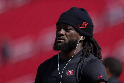FootballR - NFL - Ein Mann mit langen Dreadlocks, einer schwarzen Beanie-Mütze mit rotem „B“-Logo und einem schwarzen Nike-T-Shirt mit dem 49ers-Star Brandon Aiyuk hört über weiße Kopfhörer Musik. Der Hintergrund ist verschwommen und zeigt Rottöne. Diese Beschreibung wurde automatisch generiert.