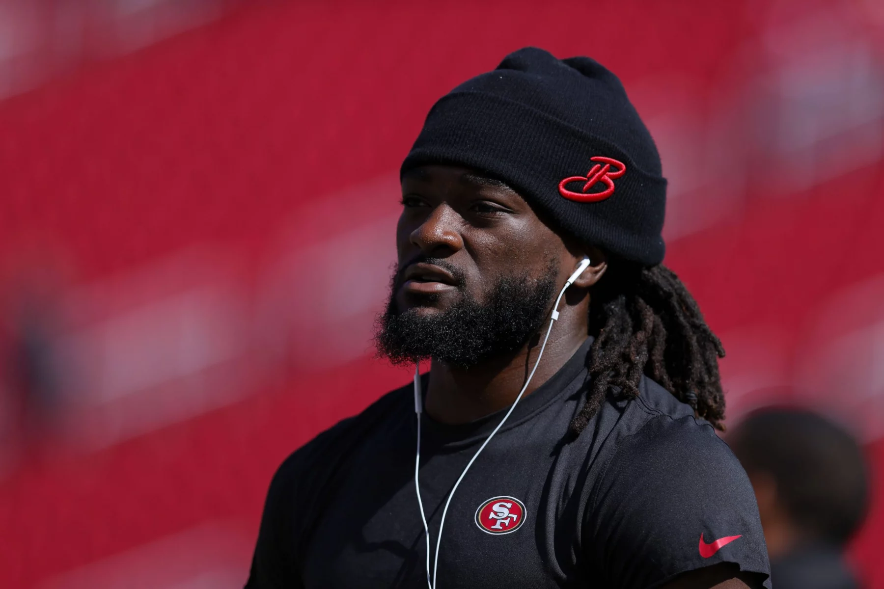 FootballR - NFL - Ein Mann mit langen Dreadlocks, einer schwarzen Beanie-Mütze mit rotem „B“-Logo und einem schwarzen Nike-T-Shirt mit dem 49ers-Star Brandon Aiyuk hört über weiße Kopfhörer Musik. Der Hintergrund ist verschwommen und zeigt Rottöne. Diese Beschreibung wurde automatisch generiert.